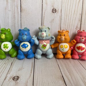 Puede incluir: Cinco figuras de oso de pl&aacute;stico alineadas. El oso verde tiene un tr&eacute;bol, el azul una nube de lluvia, el azul claro un ramo de rosas, el naranja dos girasoles y el oso rosa tiene dos corazones rojos en la parte del vientre.