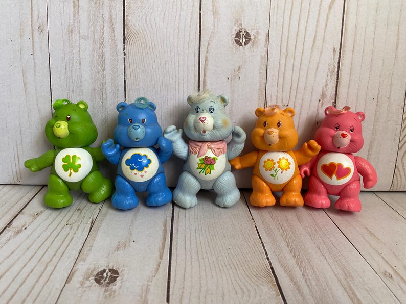 Puede incluir: Cinco figuras de oso de pl&aacute;stico alineadas. El oso verde tiene un tr&eacute;bol, el azul una nube de lluvia, el azul claro un ramo de rosas, el naranja dos girasoles y el oso rosa tiene dos corazones rojos en la parte del vientre.