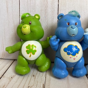 Puede incluir: Dos figuras de oso de pl&aacute;stico de pie una al lado de la otra. El oso verde presenta una insignia blanca en el vientre con un tr&eacute;bol de cuatro hojas verde. El oso azul presenta una insignia blanca en el vientre con una nube azul y corazones.