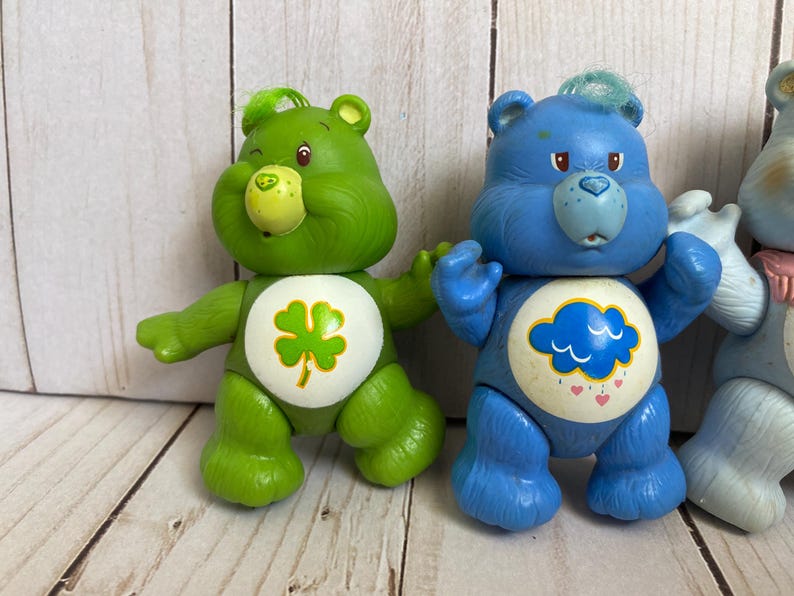 Puede incluir: Dos figuras de oso de pl&aacute;stico de pie una al lado de la otra. El oso verde presenta una insignia blanca en el vientre con un tr&eacute;bol de cuatro hojas verde. El oso azul presenta una insignia blanca en el vientre con una nube azul y corazones.