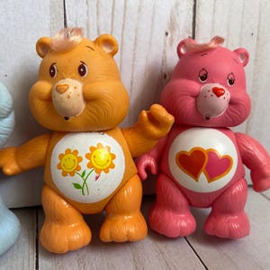 Puede incluir: Tres figuras de oso de pl&aacute;stico que incluyen un oso azul claro a la izquierda, un oso naranja con una insignia de flor amarilla en el vientre en el centro y un oso rosa con una insignia de dos corazones en el vientre a la derecha, cada uno con un mech&oacute;n de pelo.