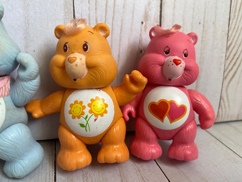 Puede incluir: Tres figuras de oso de pl&aacute;stico que incluyen un oso azul claro a la izquierda, un oso naranja con una insignia de flor amarilla en el vientre en el centro y un oso rosa con una insignia de dos corazones en el vientre a la derecha, cada uno con un mech&oacute;n de pelo.