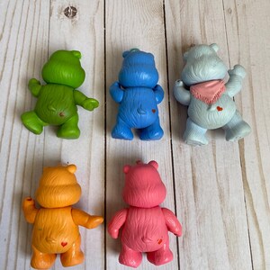 Puede incluir: Cinco peque&ntilde;as figuras de oso de pl&aacute;stico en verde, azul, azul claro, naranja y rosa. Cada oso se muestra desde atr&aacute;s, con un peque&ntilde;o s&iacute;mbolo de coraz&oacute;n rojo en la parte baja de la espalda. El oso azul claro lleva un pa&ntilde;uelo triangular rosa.