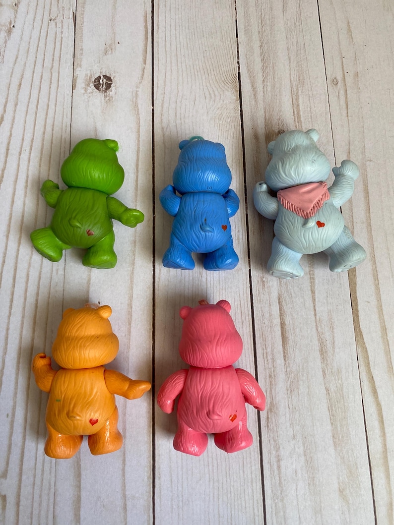 Puede incluir: Cinco peque&ntilde;as figuras de oso de pl&aacute;stico en verde, azul, azul claro, naranja y rosa. Cada oso se muestra desde atr&aacute;s, con un peque&ntilde;o s&iacute;mbolo de coraz&oacute;n rojo en la parte baja de la espalda. El oso azul claro lleva un pa&ntilde;uelo triangular rosa.