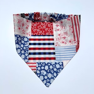 May include: A patchwork dog bandana in red, white, and blue. The bandana features various patterns, including floral, paisley, stripes, and gingham. The top edge has a gathered elastic band for a comfortable fit.