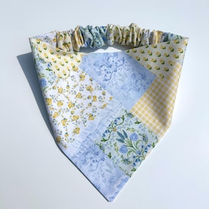 May include: A triangular fabric bandana featuring a patchwork design of various floral and gingham patterns in shades of light blue, yellow, and white. The top edge includes an elasticized, gathered band for a secure and flexible fit.