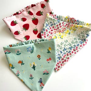 Peut inclure: Trois bandanas colorés avec différents motifs. L'un est rose clair avec des fraises rouges, un autre présente un motif floral et fruité, et le troisième est vert clair avec des fleurs, des champignons et un papillon. Chaque bandana a un bord à volants.