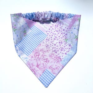 May include: A triangular pet bandana featuring a patchwork design of light purple and blue fabrics. The pattern includes floral prints, rose motifs, and a blue and white gingham check. The top edge has a gathered elastic band for a secure fit.