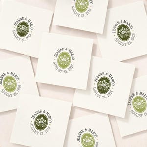 May include: Several white square cards featuring a green and white floral design and the text "JEANNE & MARCO AUGUST 23, 2026". The cards are arranged on a light surface, with the text in a circular pattern.