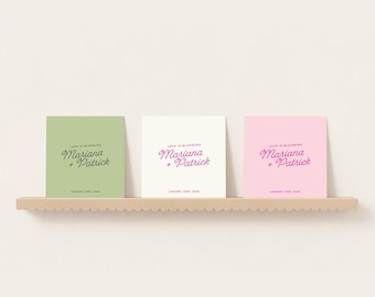 May include: Three square cards in green, white, and pink, each with the text "Love is Blooming" and "Mariana + Patrick" in a script font. The cards are displayed on a light brown shelf.