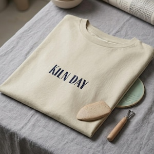 May include: A beige short-sleeved t-shirt with the text KILN DAY printed in dark blue letters on the chest. The shirt is folded and placed near a small speckled tan ceramic tool and a light green circular ceramic piece with a wooden handle.