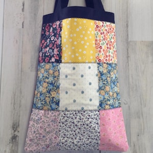 May include: A patchwork tote bag with a navy blue handle and top. The bag features a variety of floral patterns in pink, blue, yellow, and white. The bag is made of fabric and is designed for carrying items.