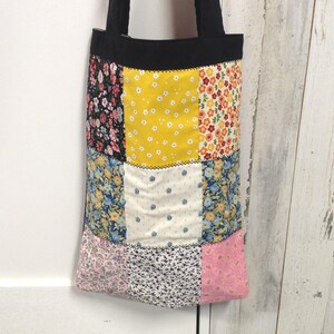 May include: A patchwork tote bag with a black top and straps. The bag features a variety of floral patterns in pink, yellow, blue, and black. The bag is made of fabric and is designed for carrying items.