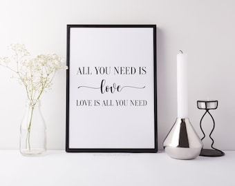 May include: Black and white framed print with the text "All you need is love. Love is all you need." The print is displayed on a white surface with a vase of white flowers and a silver candle holder.