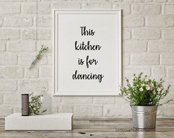 May include: White framed print with black calligraphy that reads "This kitchen is for dancing". The print is hanging on a white brick wall. There are small white flowers in a metal bucket and a glass jar on a white shelf.