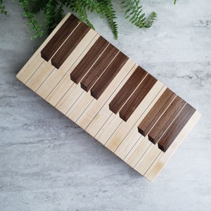 May include: A wooden keyboard-shaped cutting board. The keys alternate between light-colored wood and dark brown wood. The board is rectangular and angled, with a fern in the background.