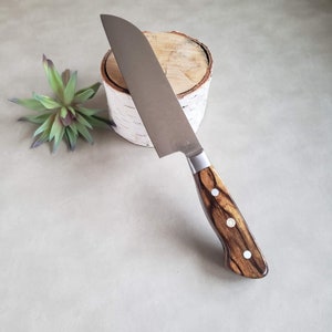 May include: A silver chef's knife with a wooden handle. The handle has a dark wood grain with three white dots. The knife is resting on a wooden log.