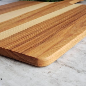Puede incluir: Una tabla de cortar de madera con un patr&oacute;n de veta de madera clara y oscura. La tabla tiene un borde redondeado y est&aacute; hecha de dos tipos diferentes de madera.