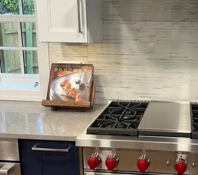 Puede incluir: Una estufa de gas de acero inoxidable con cuatro perillas rojas y una rejilla de hierro fundido negro. La estufa est&aacute; en una cocina con gabinetes blancos y un protector contra salpicaduras de azulejos blancos. Un libro de cocina est&aacute; en un soporte de madera a la izquierda de la estufa.