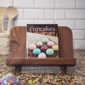 Puede incluir: Un soporte de libro de cocina de madera con un libro de cocina titulado "cupcakes...made simple" en la parte superior. La portada del libro de cocina presenta una variedad de cupcakes coloridos.