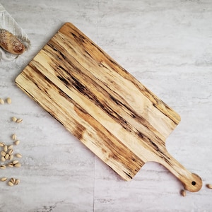May include: A large, rectangular wooden serving board with a long handle. The board has a natural wood finish with dark brown streaks and a light brown background. The handle has a round knob at the end.