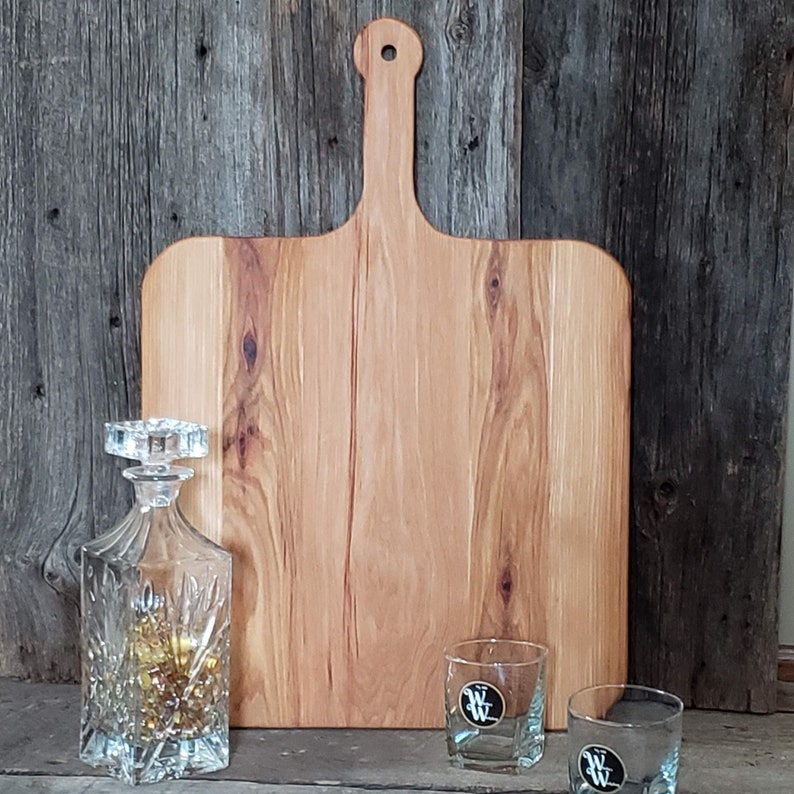 Puede incluir: Una tabla de cortar de madera grande, rectangular, con un asa. La tabla es de madera clara con un patr&oacute;n de veta natural. Est&aacute; sobre una superficie de madera r&uacute;stica.