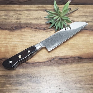 May include: A black-handled kitchen knife with a Damascus steel blade, resting on a wooden cutting board. The knife has a textured blade and a black handle with gold accents.