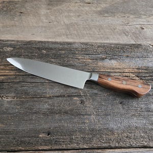 May include: A stainless steel chef's knife with a wooden handle. The blade is silver and the handle is a dark brown with a decorative emblem. The knife is set against a rustic wooden background.