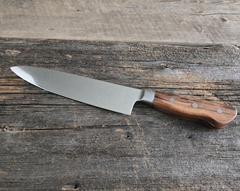 May include: A stainless steel chef's knife with a wooden handle. The blade is silver and the handle is a dark brown with a decorative emblem. The knife is set against a rustic wooden background.