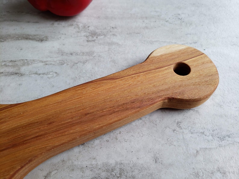 Puede incluir: Un utensilio de cocina de madera con un mango largo y una cabeza redondeada con un agujero en el centro. El utensilio est&aacute; hecho de madera marr&oacute;n claro con un patr&oacute;n de veta natural.