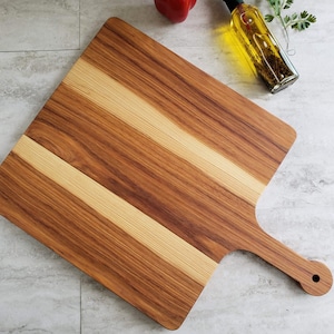 Puede incluir: Una tabla de cortar de madera grande, rectangular, con un mango. La tabla est&aacute; hecha de dos tipos diferentes de madera, creando un patr&oacute;n a rayas. La tabla es de color marr&oacute;n claro y tiene un acabado natural.