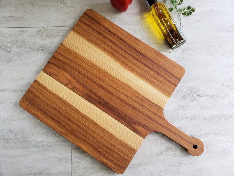 Puede incluir: Una tabla de cortar de madera grande, rectangular, con un mango. La tabla est&aacute; hecha de dos tipos diferentes de madera, creando un patr&oacute;n a rayas. La tabla es de color marr&oacute;n claro y tiene un acabado natural.