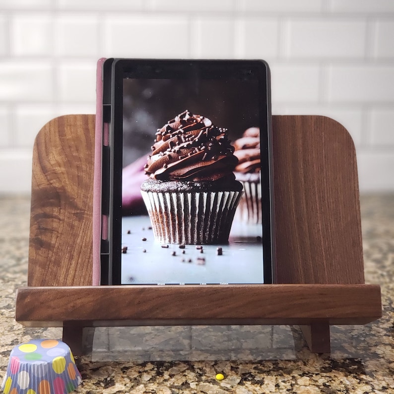 Puede incluir: Un soporte de madera con un acabado marr&oacute;n oscuro sostiene una tableta que muestra una foto de un cupcake de chocolate con chispas. El soporte tiene un borde en la parte delantera y trasera para sujetar la tableta en su lugar.