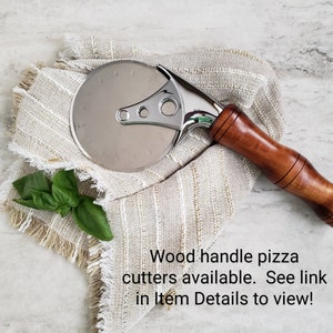 Puede incluir: Un cortador de pizza plateado con mango de madera. El cortador est&aacute; sobre una tela beige con borde de flecos.  Cortadores de pizza con mango de madera disponibles. &iexcl;Ver enlace en Detalles del art&iacute;culo para ver!