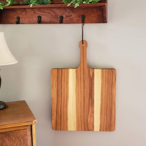 Puede incluir: Una tabla de cortar cuadrada de madera con un asa cuelga de un gancho de pared. La tabla est&aacute; hecha de madera clara y oscura, creando un patr&oacute;n a rayas.