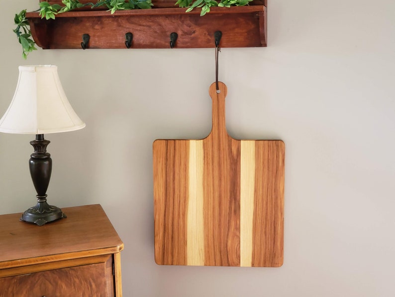 Puede incluir: Una tabla de cortar cuadrada de madera con un asa cuelga de un gancho de pared. La tabla est&aacute; hecha de madera clara y oscura, creando un patr&oacute;n a rayas.