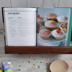 Puede incluir: Un libro de cocina abierto en una receta de cupcakes glaseados. La receta incluye ingredientes e instrucciones. La imagen muestra un primer plano del libro con un soporte de libro de madera que lo mantiene abierto. Hay chispas de colores y una cuchara de madera en la mesa.