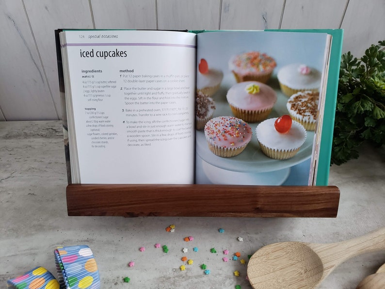 Puede incluir: Un libro de cocina abierto en una receta de cupcakes glaseados. La receta incluye ingredientes e instrucciones. La imagen muestra un primer plano del libro con un soporte de libro de madera que lo mantiene abierto. Hay chispas de colores y una cuchara de madera en la mesa.