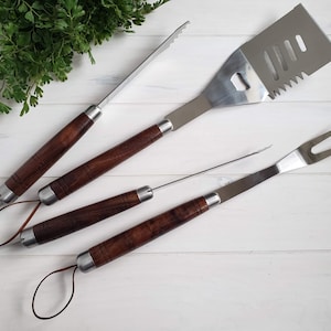 May include: A set of three stainless steel grilling tools with wooden handles. The tools include a spatula, tongs, and a fork.