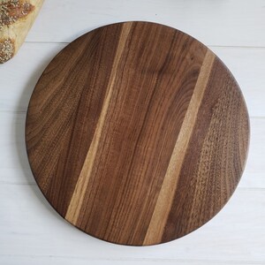 May include: A circular wooden serving board crafted from dark brown walnut wood with visible natural grain patterns. The board features a smooth, flat surface with distinct light and dark wood tones running vertically across the center.