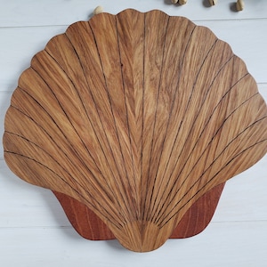 May include: A wooden seashell-shaped serving tray with a natural wood finish and a reddish-brown interior. The tray is carved with a detailed design of lines radiating from the center.