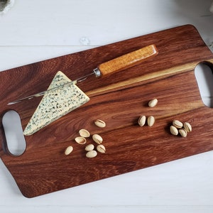 May include: A wooden cutting board with two handles, a wedge of blue cheese, a small knife, and pistachios on top.