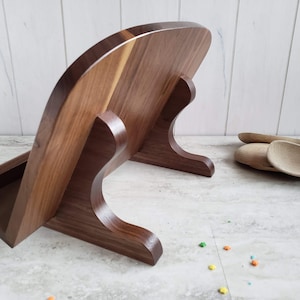 Puede incluir: Un soporte para cucharas de madera de nogal con una parte posterior curva y un peque&ntilde;o estante para guardar cucharas. El soporte est&aacute; sobre una encimera blanca.
