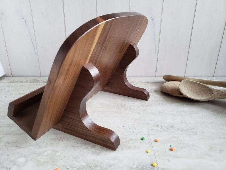 Puede incluir: Un soporte para cucharas de madera de nogal con una parte posterior curva y un peque&ntilde;o estante para guardar cucharas. El soporte est&aacute; sobre una encimera blanca.