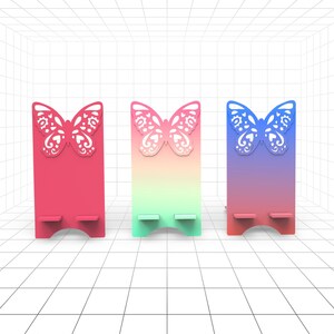 以下が含まれることがあります： 蝶のデザインの携帯電話スタンドが3つあります。スタンドは木でできており、グラデーションカラーになっています。最初のスタンドはピンク、2番目はグリーン、3番目はブルーです。