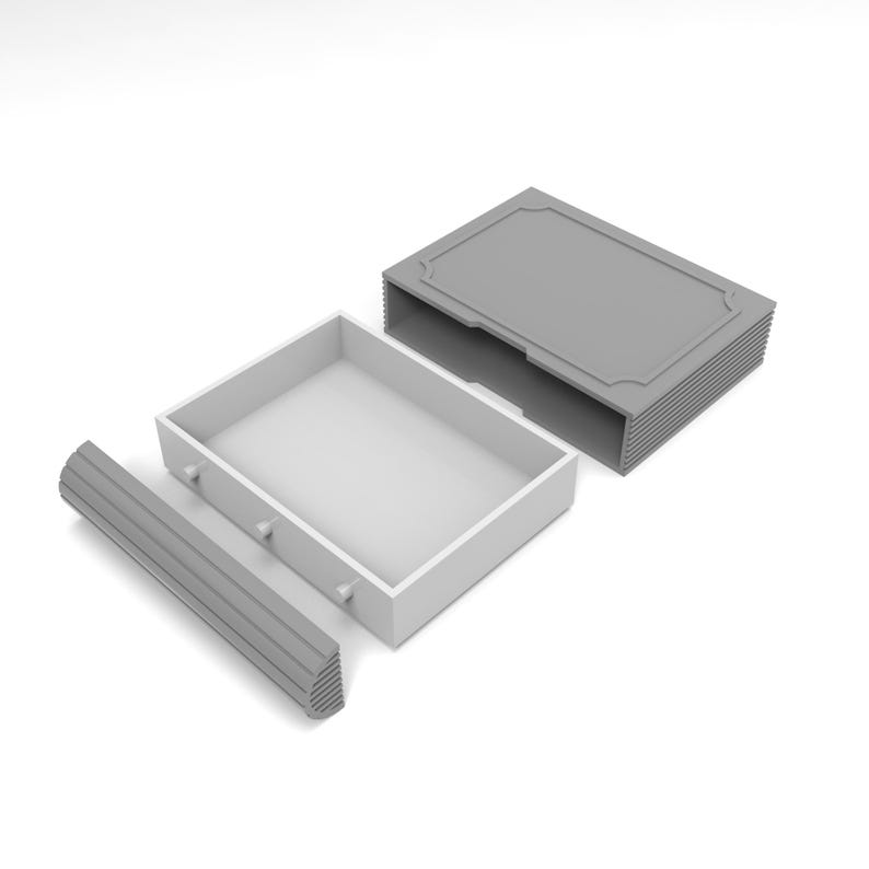 Puede incluir: Caja bicolor abierta, con tapa gris e interior blanco. La caja es rectangular con un borde decorativo en la tapa. Una pieza gris y estriada est&aacute; a la izquierda. Dos peque&ntilde;os botones son visibles en la caja.