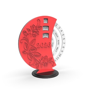 May include: A red decorative desk calendar for the year 2026. The calendar features a floral design and displays the month of January and the day 30. The days of the week and months are displayed on a rotating wheel.