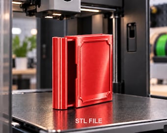 以下が含まれることがあります： 本のようなデザインの鮮やかな赤色の3Dプリント製収納ボックス。隆起した縁を持つ長方形のフレームとリブ状の背表紙が特徴で、プリンターのベッドの上に置かれています。オブジェクトの下の表面には、白色でSTL FILEという文字が印字されています。