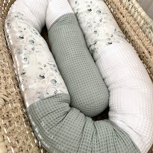 Op de afbeelding: Een babynest in een rieten mand. Het nest combineert witte wafelstof, saliegroene wafelstof en stof bedrukt met een botanisch bladerpatroon in grijstinten en groen. Het nest is ontworpen voor baby's.
