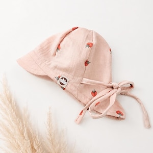 Könnte beinhalten: Eine hellrosa Haube mit einem wiederkehrenden Muster aus kleinen roten Erdbeeren mit grünen Blättern. Die Haube hat eine breite Krempe und lange Stoffbänder an der Seite zum Binden. Ein kleines rechteckiges Etikett ist an der Krempe befestigt.